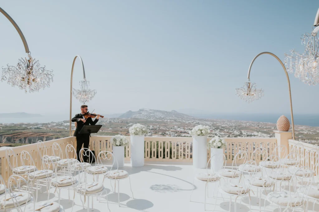 Hazel & Soltan wedding at Madame Sousou, Santorini — the terrace set for the ceremony, a musician playing beside the chandelier arches